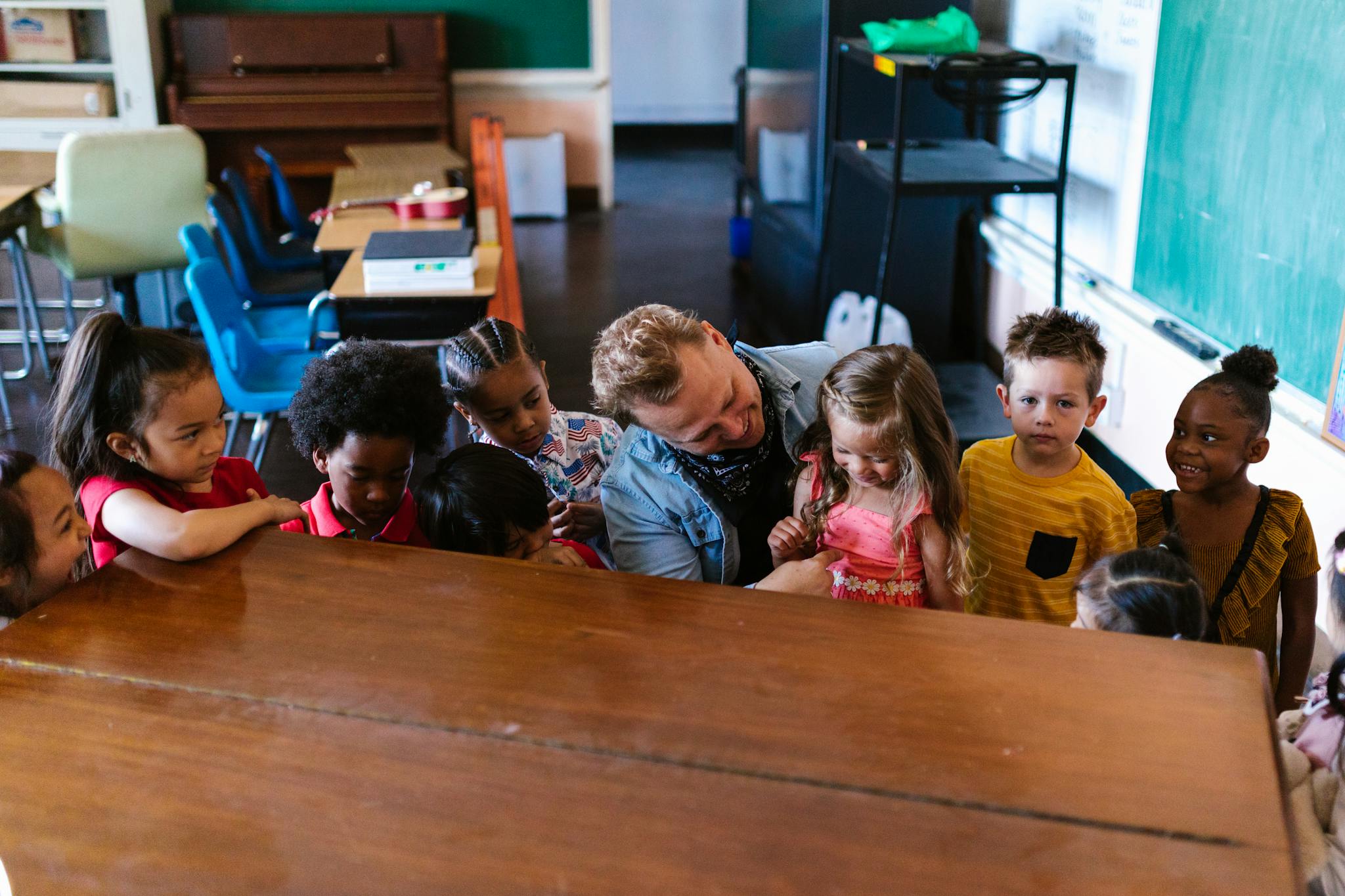 A warm classroom scene showing a teacher interacting with diverse preschool children around a piano.